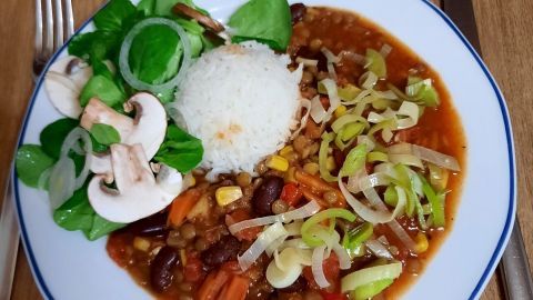 Eine bunte Chili sin Carne mit Linsen und Reis liegt auf einem weißen Teller, umgeben von einem frischen Salat.