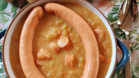 Eine Schüssel mit cremiger Kartoffelsuppe, garniert mit zwei Würstchen, steht neben frischen Kartoffeln auf einem Tisch.