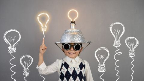 Ein Kind trägt einen Aluhut und Sonnenbrillen, hält eine leuchtende Glühbirne hoch, umgeben von gezeichneten Glühbirnen, die Ideen symbolisieren.