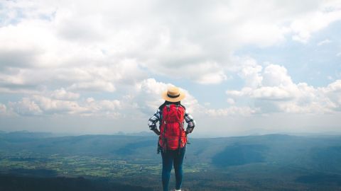 Eine Person mit rotem Rucksack und Strohhut steht auf einem Felsen und genießt die Aussicht auf eine weite Landschaft.