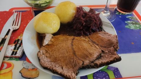 Zarter Rinderbraten liegt auf einem Teller, umgeben von Kartoffelknödeln und Rotkraut, mit einem Salat und einem Getränk.