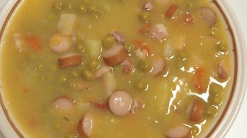Eine Schüssel Erbsensuppe mit Würstchen schwimmt in einer sämigen Brühe, auf einem Tisch mit dezentem Muster.