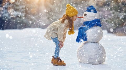 Ein Mädchen mit einer gelben Mütze beugt sich neugierig zu einem Schneemann im winterlichen Schnee, während es schneit.