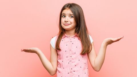 Ein Mädchen in einem pinken Shirt zuckt mit den Schultern und sieht verwirrt aus, vor einem rosafarbenen Hintergrund.