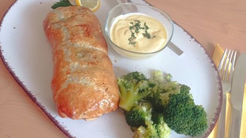 Ein knuspriger Blätterteig mit Lachs und Spinat liegt auf einem Teller, ergänzt von Brokkoli und einer Sauce.