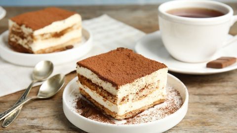 Eine Portion klassisches Tiramisu liegt auf einem weißen Teller, während eine Tasse Tee im Hintergrund steht.
