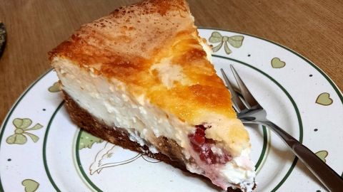 Ein Stück Käsekuchen mit Himbeeren liegt auf einem gemusterten Teller und wird von einer Gabel begleitet.