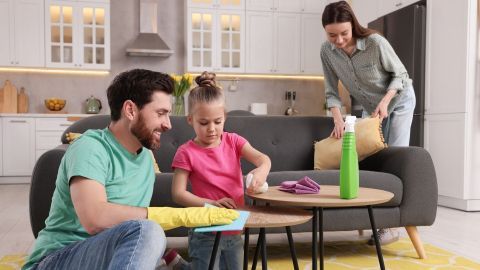 Ein Vater mit gelben Handschuhen hilft seiner Tochter, während sie den Tisch mit einem Tuch reinigen; die Mutter räumt im Hintergrund auf.