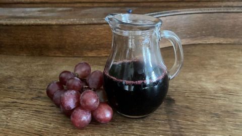 Ein Glasbehälter steht auf einem Holztisch, gefüllt mit tiefrotem Balsamico Essig, neben frischen roten Trauben.