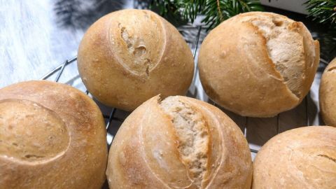 Dinkel-Sauerteigbrötchen liegen frisch gebacken auf einem Gitter, umgeben von Tannenzweigen im Hintergrund.