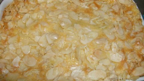 Ein goldbraun gebackener Ofenschlupfer mit mandel-ähnlicher Oberseite liegt in einer weißen Auflaufform auf einem Holzbrett.
