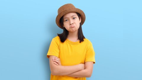 Ein Mädchen in einem gelben T-Shirt mit verschränkten Armen schaut unzufrieden und trägt einen Strohhut vor einem blauen Hintergrund.