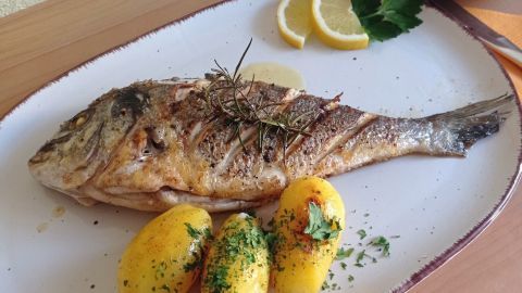 Eine gebratene Dorade liegt auf einem Teller, umgeben von Zitronenscheiben und Kräutern, neben kleinen Kartoffeln.