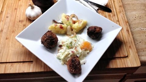 Auf einem weißen Teller liegen bräunliche Hackfleischbällchen, umgeben von Kartoffeln, Krautsalat und einer Knoblauchzehe.