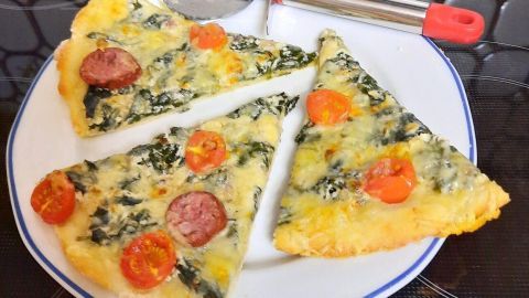 Drei Stücke Pizza mit Spinat und Tomaten liegen auf einem Teller, daneben ein Pizzaschneider auf einer modernen Oberfläche.
