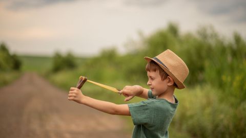 Ein Junge mit einem Strohhut spielt mit einer selbstgebauten Schleuder auf einem ruhigen Landweg, umgeben von Bäumen.