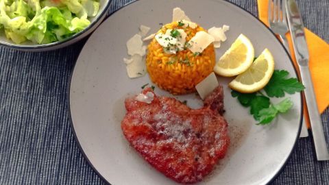 Ein knuspriges Kotelett liegt neben einem gelben Safranrisotto, ergänzt durch Zitrone und frischen Kräutern auf einem Teller.
