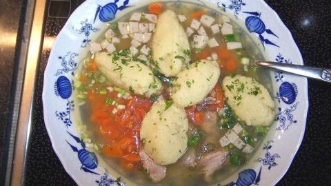 Eine gefüllte Schüssel Geflügelsuppe mit Grießnockerl schimmert klar, umgeben von Gemüse und Kräutern.