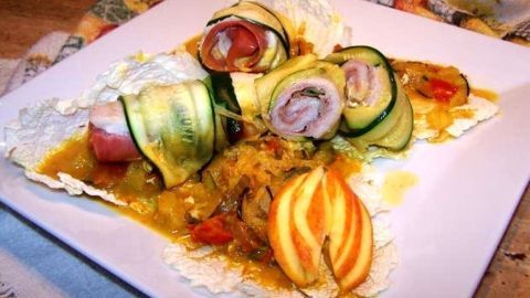 Ein kunstvoll angerichteter Teller zeigt Röllchen aus Zucchini mit Schinken, umgeben von einer bunten Gemüsesauce und Apfelstücken.
