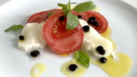 Eine elegante Anrichtung mit Tomatenscheiben, frischem Basilikum und Balsamico-Perlen auf einem weißen Teller.