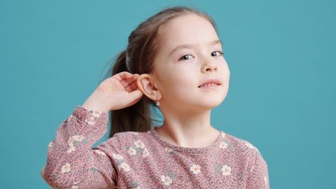 Ein kleines Mädchen mit langen Haaren hält die Hand ans Ohr und schaut neugierig, während sie vor einem blauen Hintergrund steht.