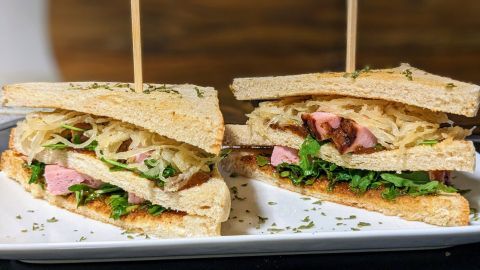 Ein doppelt geschichtetes Leberkäse-Sandwich mit Sauerkraut und Rucola auf einem weißen Teller in einem modernen Ambiente.