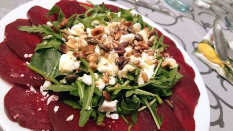 Rote Bete Carpaccio wird elegant auf einem Teller präsentiert, umgeben von Rucola, Feta und gerösteten Sonnenblumenkernen.