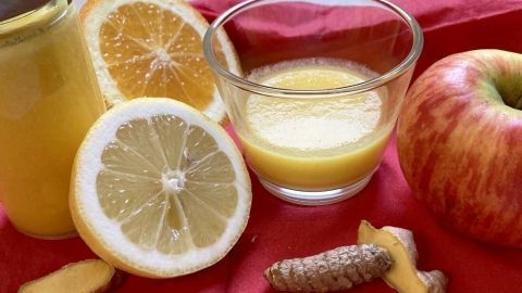 Ein gelbes Getränk in einer Glastasse steht neben frischen Zutaten wie Ingwer, Zitrone und einem Apfel auf rotem Untergrund.
