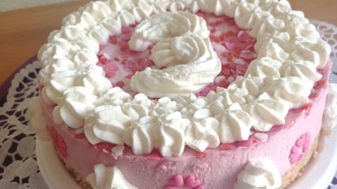 Eine rosa Kirschtorte mit Sahne und Blümchen steht auf einem filigranen Teller, umgeben von Holzoberfläche.