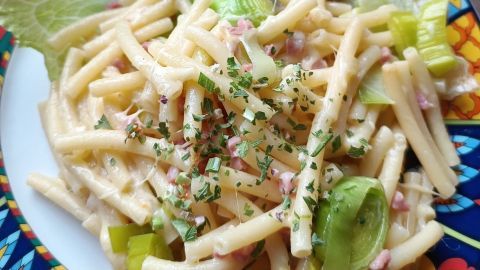 Eine cremige Nudelpfanne mit Speck, garniert mit frischen Kräutern, serviert auf buntem Teller neben Salat und Kirschtomaten.