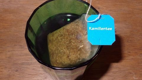 Eine Tasse Kamillentee steht auf einem Tisch, während der Teebeutel im Wasser zieht. Der Tee ist leicht bräunlich.