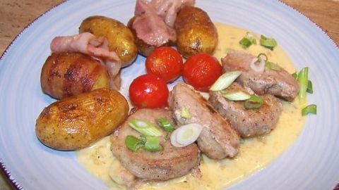 Zartes Schweinelendchen liegt auf einer Sauce, umgeben von knusprigen Speckkartoffeln und Kirschtomaten auf einem Teller.