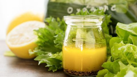 Ein Glas mit Zitronen-Salatsoße steht auf einem Holzuntergrund, umgeben von frischem Salat und Zitronen.