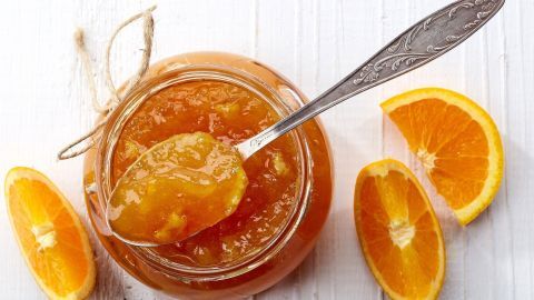 Ein Glas Orangenmarmelade steht auf einem weißen Tisch, während ein Löffel eine Portion daraus entnimmt, umgeben von frisch geschnittenen Orangen.
