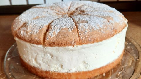 Eine hohe Käsesahnetorte mit Biskuitboden steht auf einem gläsernen Teller in einer gemütlichen Küche.
