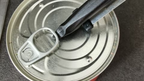 Ein Dosenöffner hebt den Deckel einer roten Konservendose auf einer dunklen Arbeitsfläche, um die darin enthaltene Speise freizulegen.
