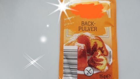 Eine Packung Backpulver liegt auf einer hellen Oberfläche, umgeben von Glanzlichtern, und zeigt einen Kuchen.