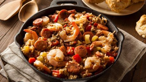Ein großer, runder Topf mit Jambalaya, gefüllt mit Garnelen, Wurst, Gemüse und Reis, steht auf einem Tisch neben Brot.