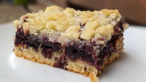 Ein Stück Kirschstreuselkuchen mit krümeliger Streuselschicht liegt auf einem weißen Teller, umgeben von unscharfem Hintergrund.