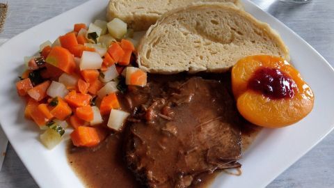 Ein Teller mit Sauerbraten, umgeben von Gemüse, Brot und einer Pfirsichhälfte, serviert auf einem weißen Teller.