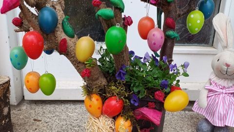 Bunt dekoriertes Ostergesteck mit Eieranhängern und Blumen steht vor einer Haustür auf dem Boden.