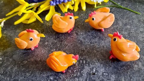 Bunte Fimo-Küken stehen fröhlich auf einem schwarzen Tisch, umgeben von gelben und blauen Blumen, passend zu Ostern.