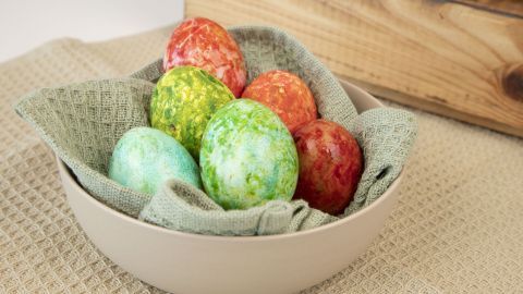 Bunte Eier liegen in einer Schale, die mit einem Tuch ausgelegt ist, und symbolisieren Traditionen zu Ostern.