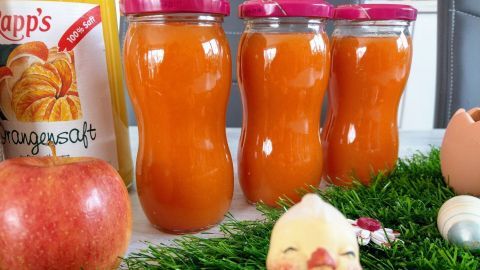 Glasbehälter mit leuchtend orangefarbener Karotten-Apfel-Konfitüre stehen auf einem Tisch, umgeben von Dekoration und einem Apfel.