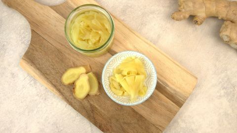 Eingelegter Ingwer liegt in einer Schale auf einem Holzbrett neben einem Glas, umgeben von frischem Ingwer.