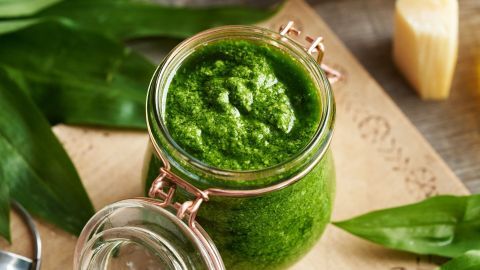 Ein Glas mit grünem Haselnuss-Pesto steht auf einem Holzbrett, umgeben von frischem Bärlauch und Käse.