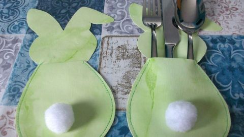 Zwei grüne Bestecktaschen in Hasenoptik halten Besteck und sind dekorativ auf einem bunten Tischmuster platziert.