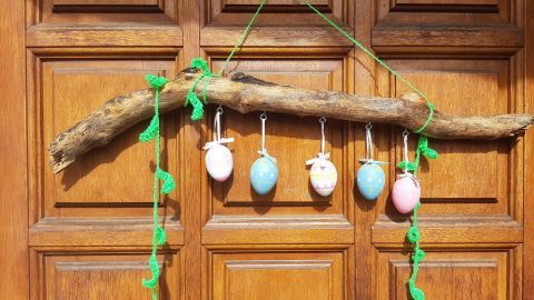 Ein schönes Holzstück hängt an einer Tür, dekoriert mit bunten Ostereiern und grünen Fäden, um Frühlingsstimmung zu verbreiten.