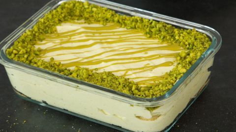 Ein schmackhaftes Pistazien-Tiramisu in einer Glasschale liegt auf einem dunklen Tisch, umgeben von einem Holzlöffel und Geschirr.