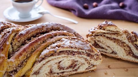 Der Hefezopf mit Nussfüllung liegt appetitlich auf einem Holztisch, neben einer Tasse Kaffee und einem lila Tuch.